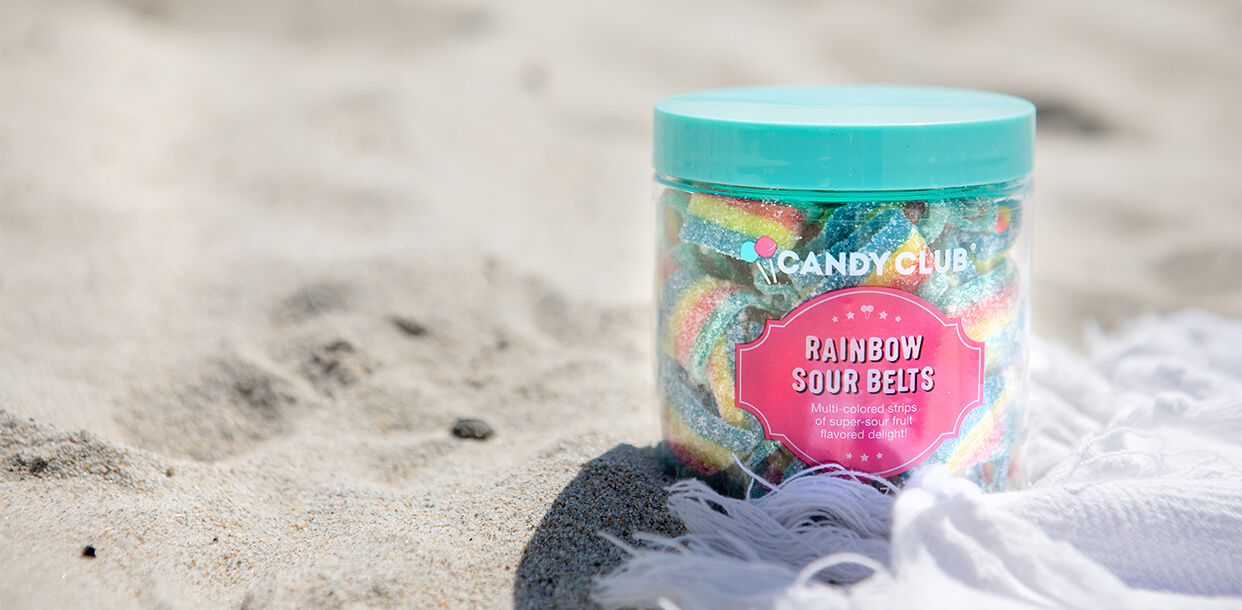 A premium cup of Candy Club Rainbow Sour Belts on the beach next to a towel on a bright summer day