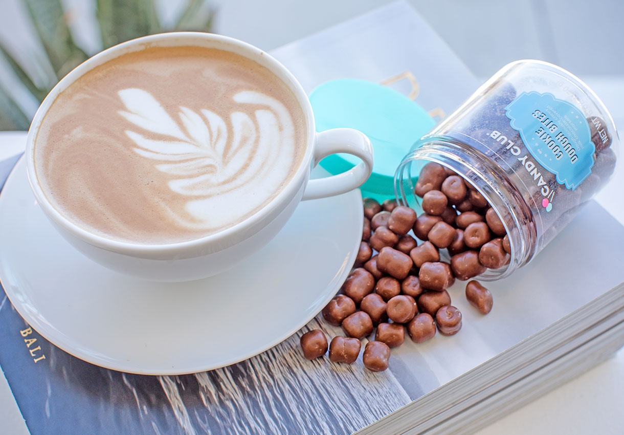 A cappuccino served next to an open cup of premium chocolate bites from Candy Club.